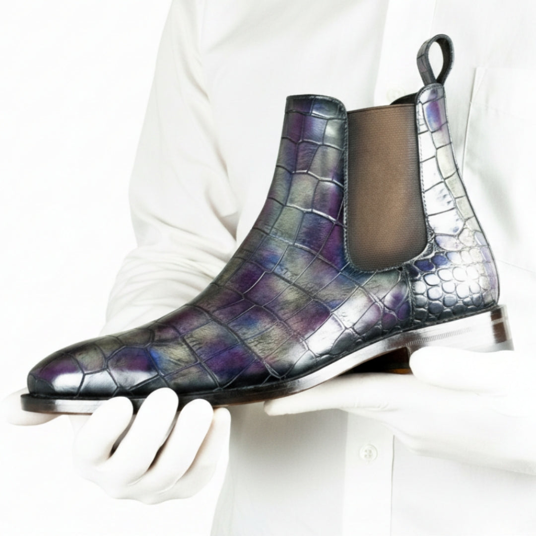 Purple crocodile-patterned Chelsea boot held by a gloved hand against a white background