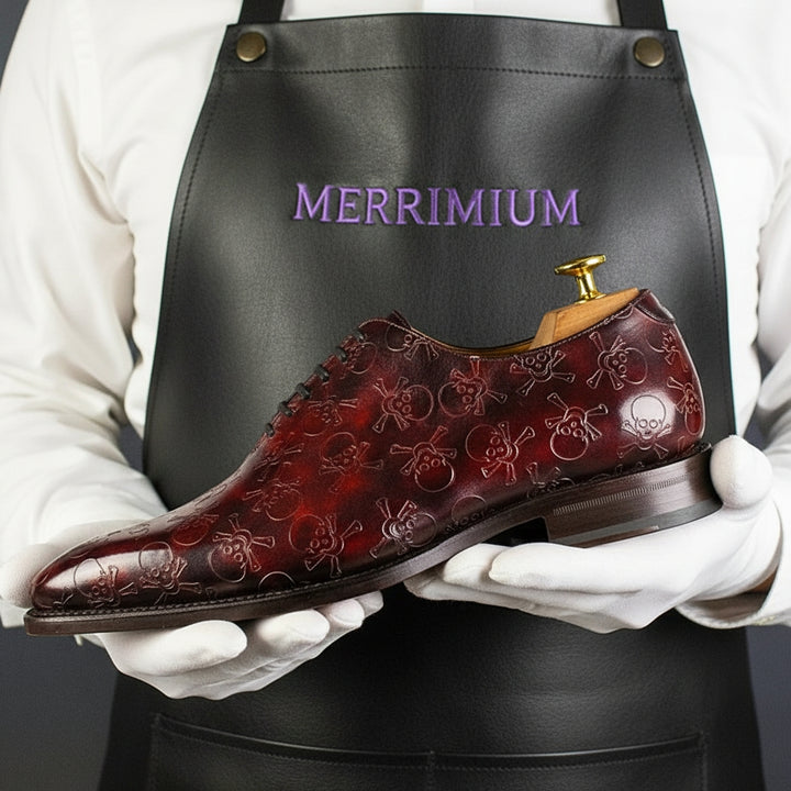 Brown leather shoe with skull patterns held by a person wearing a black apron with 'MERRIMIUM' branding.